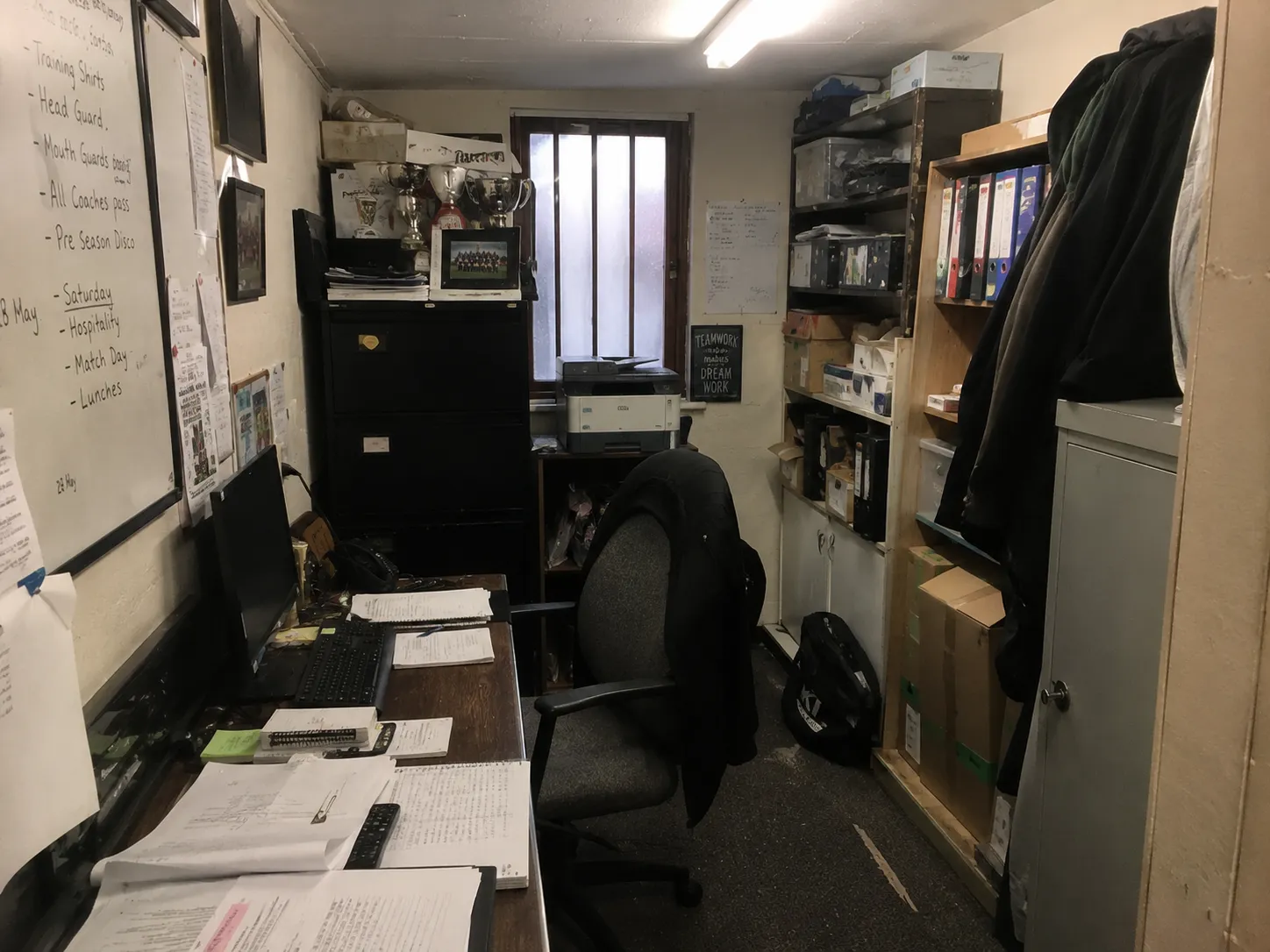 A cramped club office: a whiteboard covered in handwritten job lists, ring binders stacked on shelves, loose paperwork across the desk beside a keyboard and monitor.