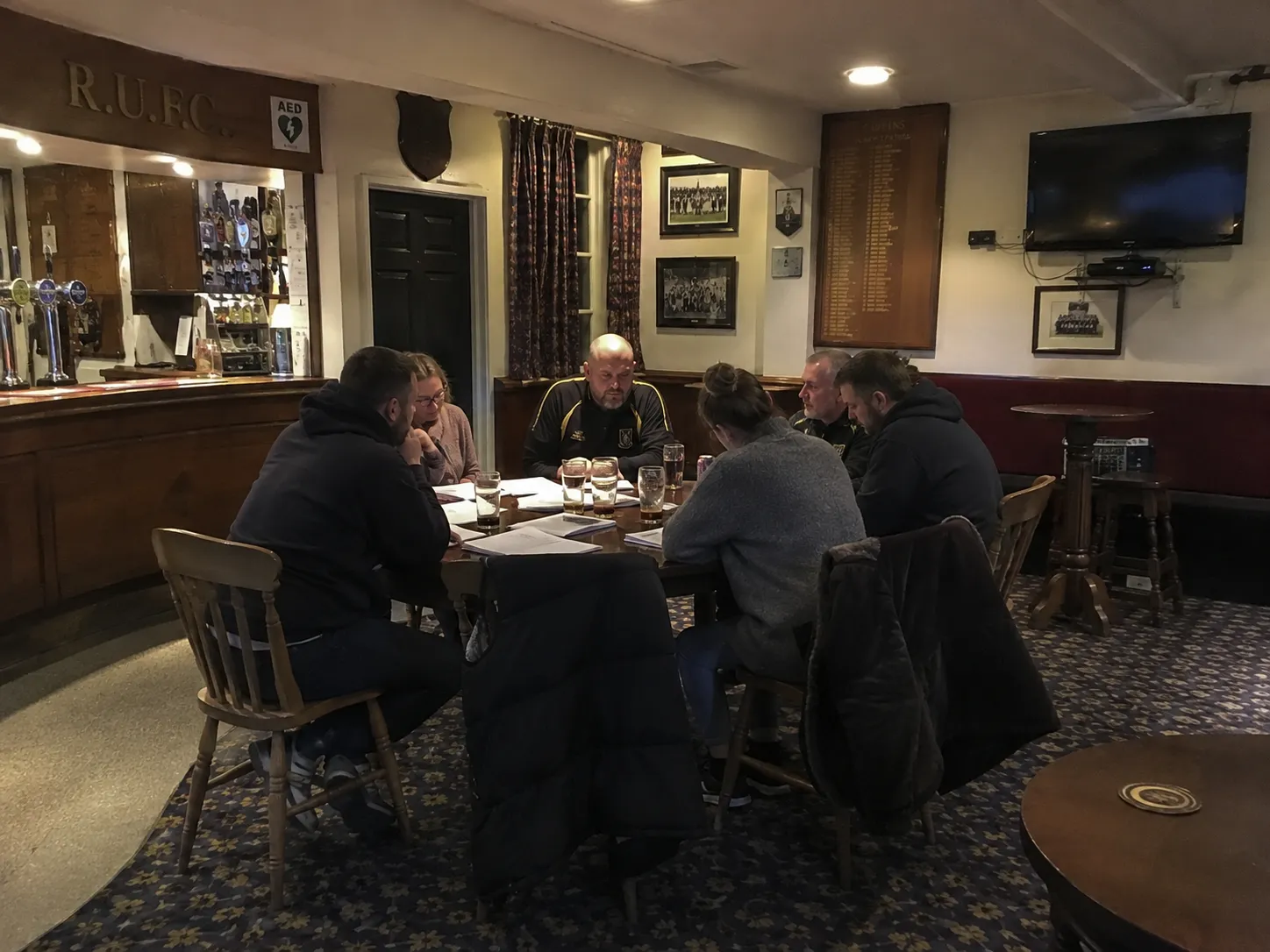 A club committee meeting around a table in the clubhouse bar, papers and pints in front of them, honours boards on the wall behind.