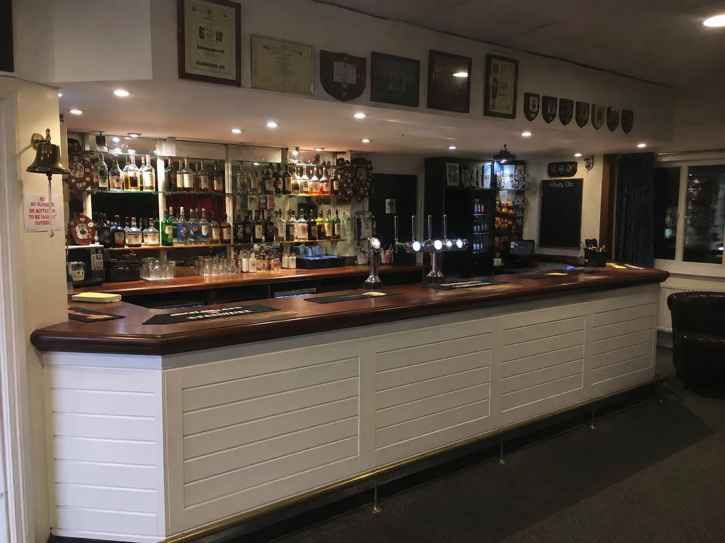 The bar in a rugby clubhouse, empty before opening, with honours shields and framed certificates above the counter and a What's On board at the end.