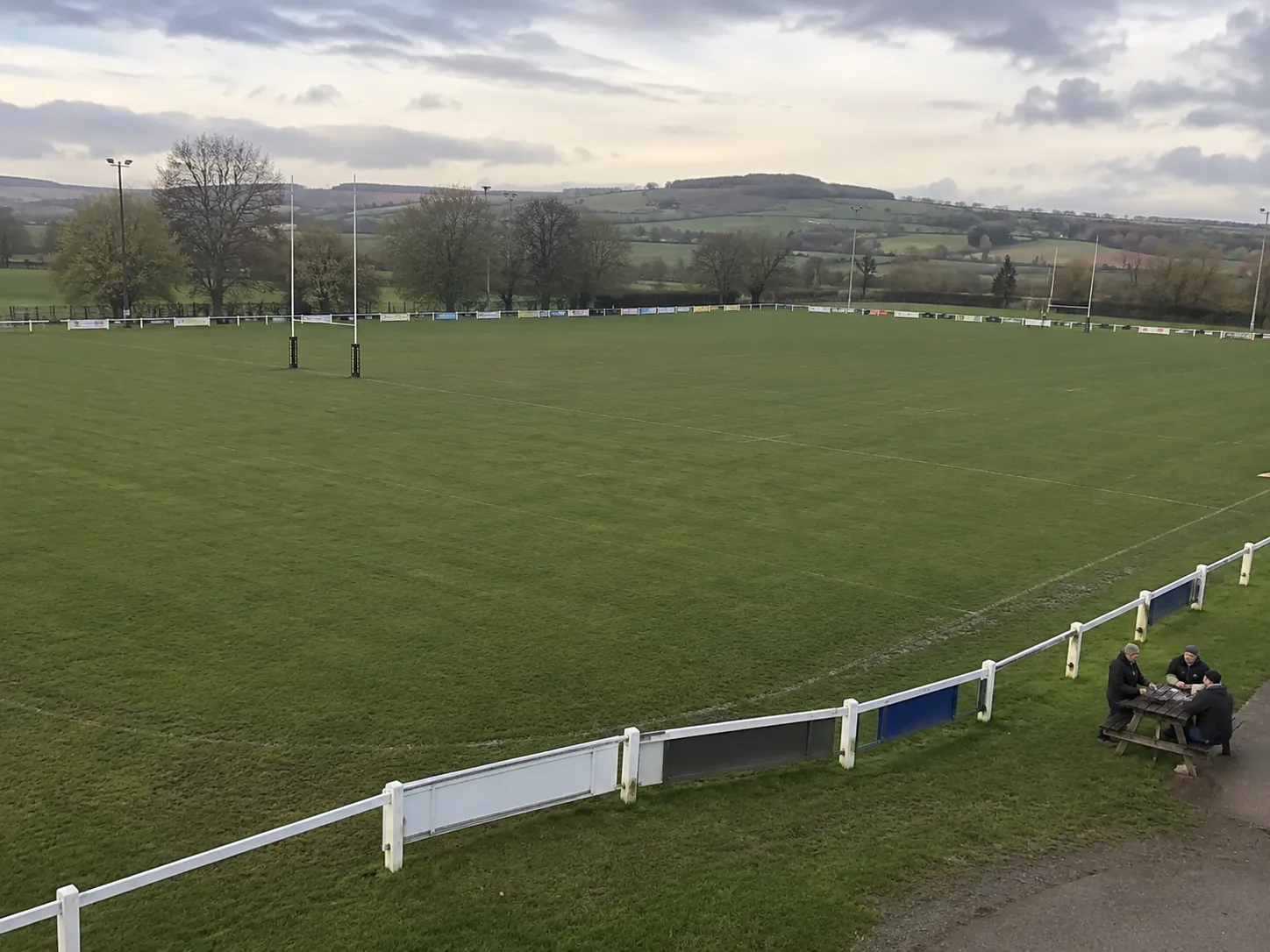 A rugby club pitch on an overcast afternoon, posts and advertising hoardings around the touchline, with three people sitting at a picnic table beside the rail.