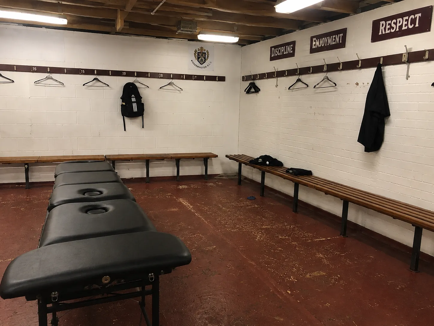 An empty club dressing room: wooden benches, coat hooks with a few hangers, and painted signs on the wall reading Discipline, Enjoyment and Respect.