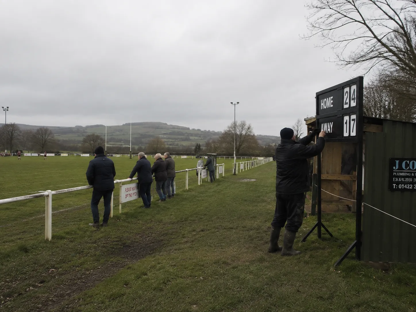 A volunteer in wellingtons reaching up to change the numbers on a hand-operated scoreboard beside the pitch, spectators watching from the rail.