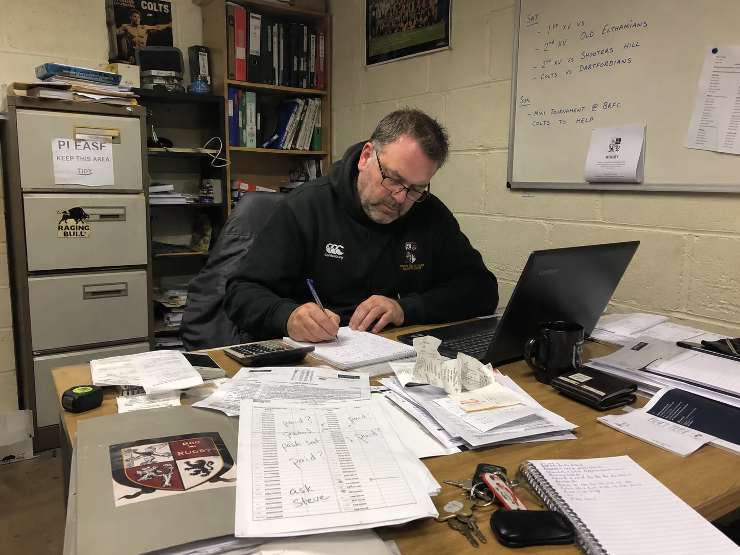 A club secretary at a paper-covered desk, annotating a printed member list by hand. Receipts, a calculator, keys and a laptop surround him; a whiteboard of Saturday fixtures is on the wall behind.