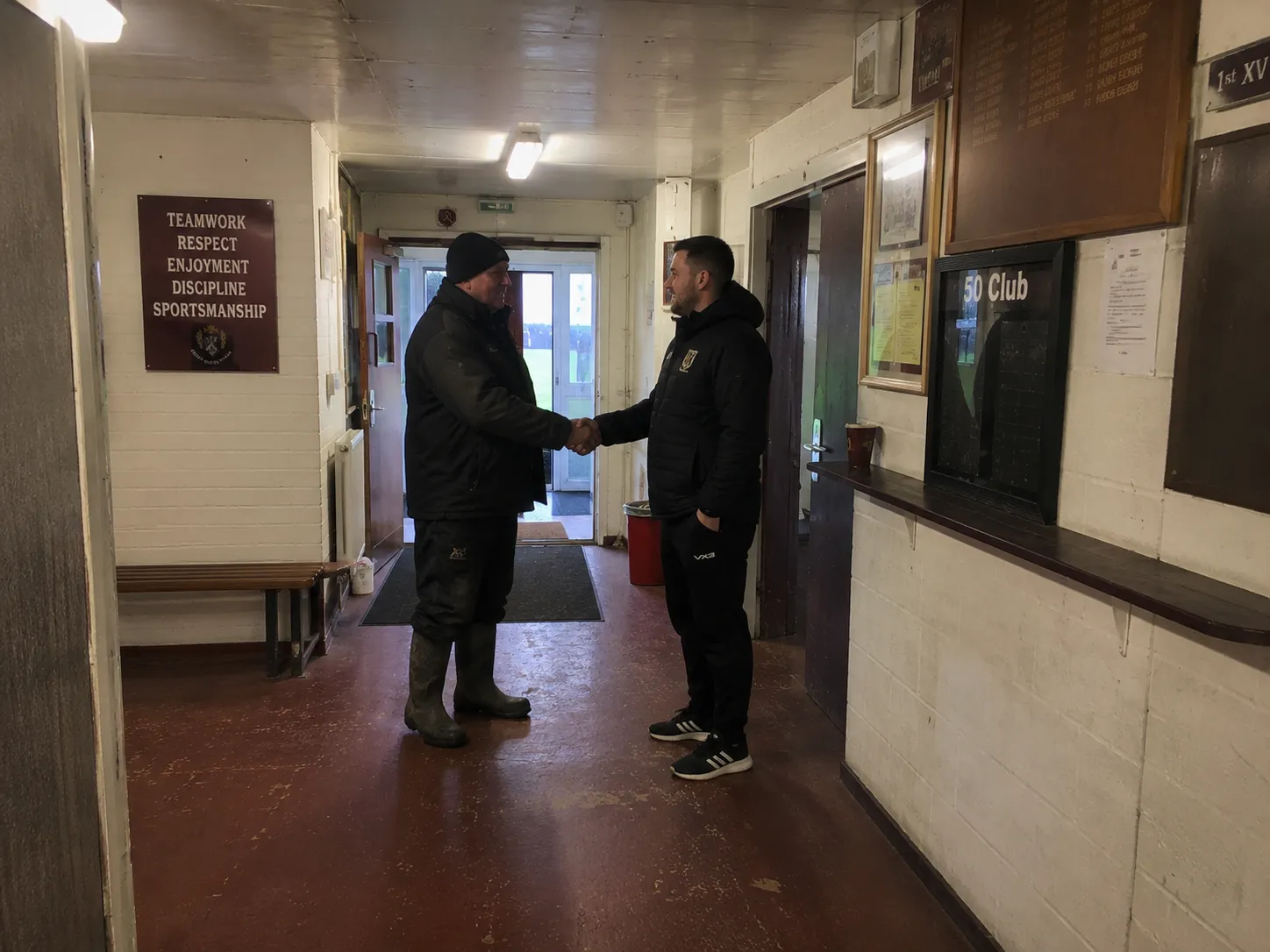 Two men shaking hands in a rugby clubhouse corridor, beneath honours boards and a sign reading Teamwork, Respect, Enjoyment, Discipline, Sportsmanship.